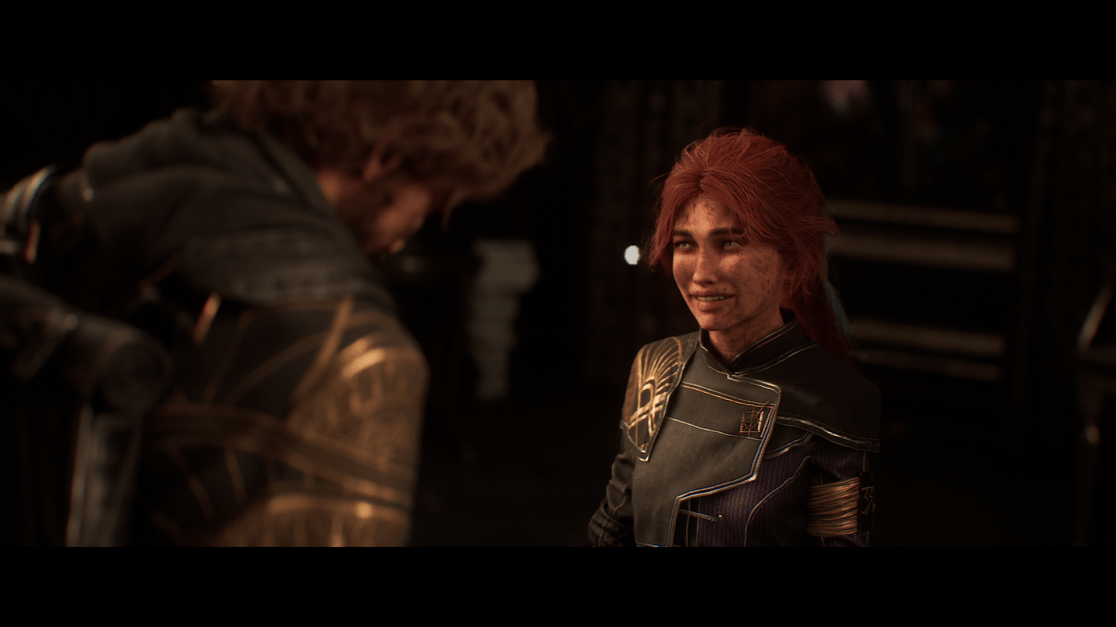 A woman with red hair and light eyes is smiling in dim lighting. There are specks of dirt on her face. She wears a dark jacket with gold trim. Part of another person, with curly brown hair and a patterned black and gold jacket, is visible on the left side.